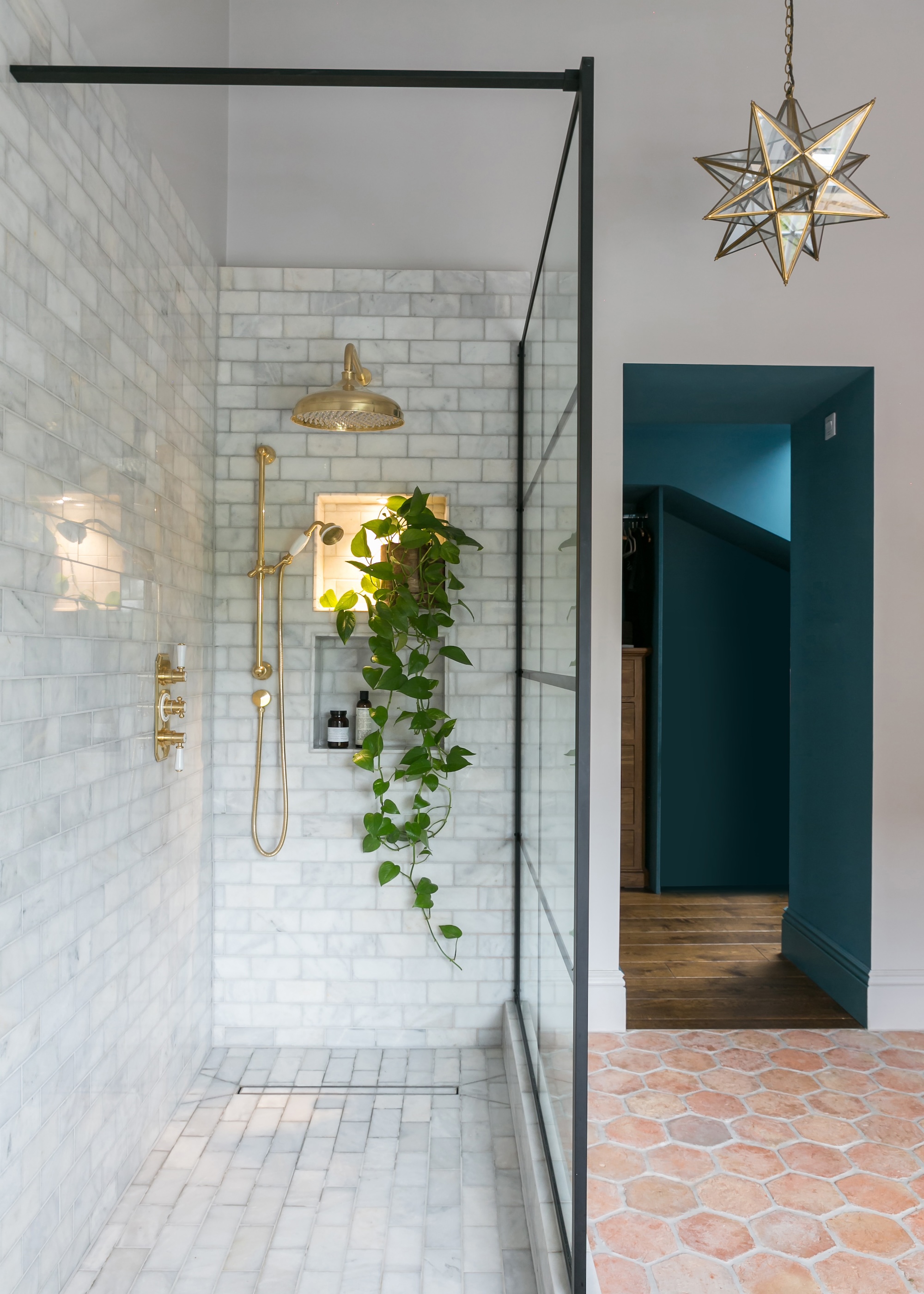 a walk in shower with neutral tiles a black grid shower screen and a light-up shower niche. Shown with a statement pendant light and terracotta floor tiles