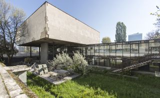 Daytime outside image of the museum, overgrown grass, concrete paved pathway, concrete level of the building, glass fronted corridors, steps, trees, blue sky