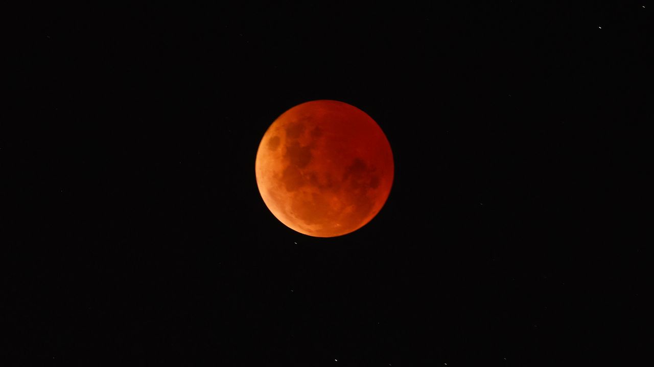A blood red moon is at the center of the image against a black sky with some stars present.