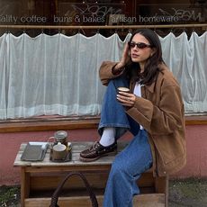 tia drinks coffee at cafe while wearing blue jeans, brown loafers and a suede jacket
