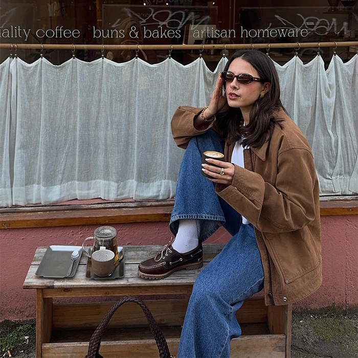 tia drinks coffee at cafe while wearing blue jeans, brown loafers and a suede jacket