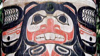 A large black, red and white wood carved totem pole shows a large face with black eyes and white teeth with feathers on either side. 