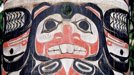 A large black, red and white wood carved totem pole shows a large face with black eyes and white teeth with feathers on either side. 