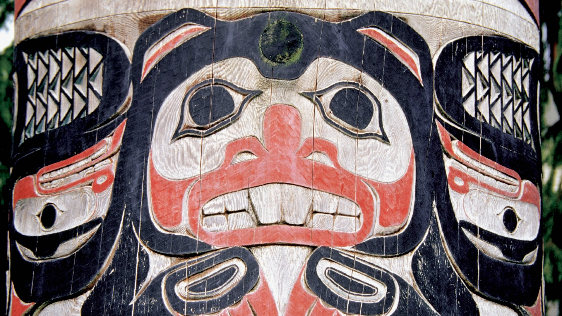 A large black, red and white wood carved totem pole shows a large face with black eyes and white teeth with feathers on either side. 