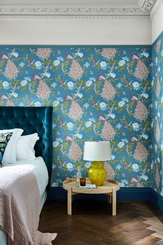 bedroom with peacock blue wallpaper, white picture rail and ceiling, wooden floors, blue skirting, wooden round side table, yellow based lamp, blue upholstered bed, white bedding, pink throw