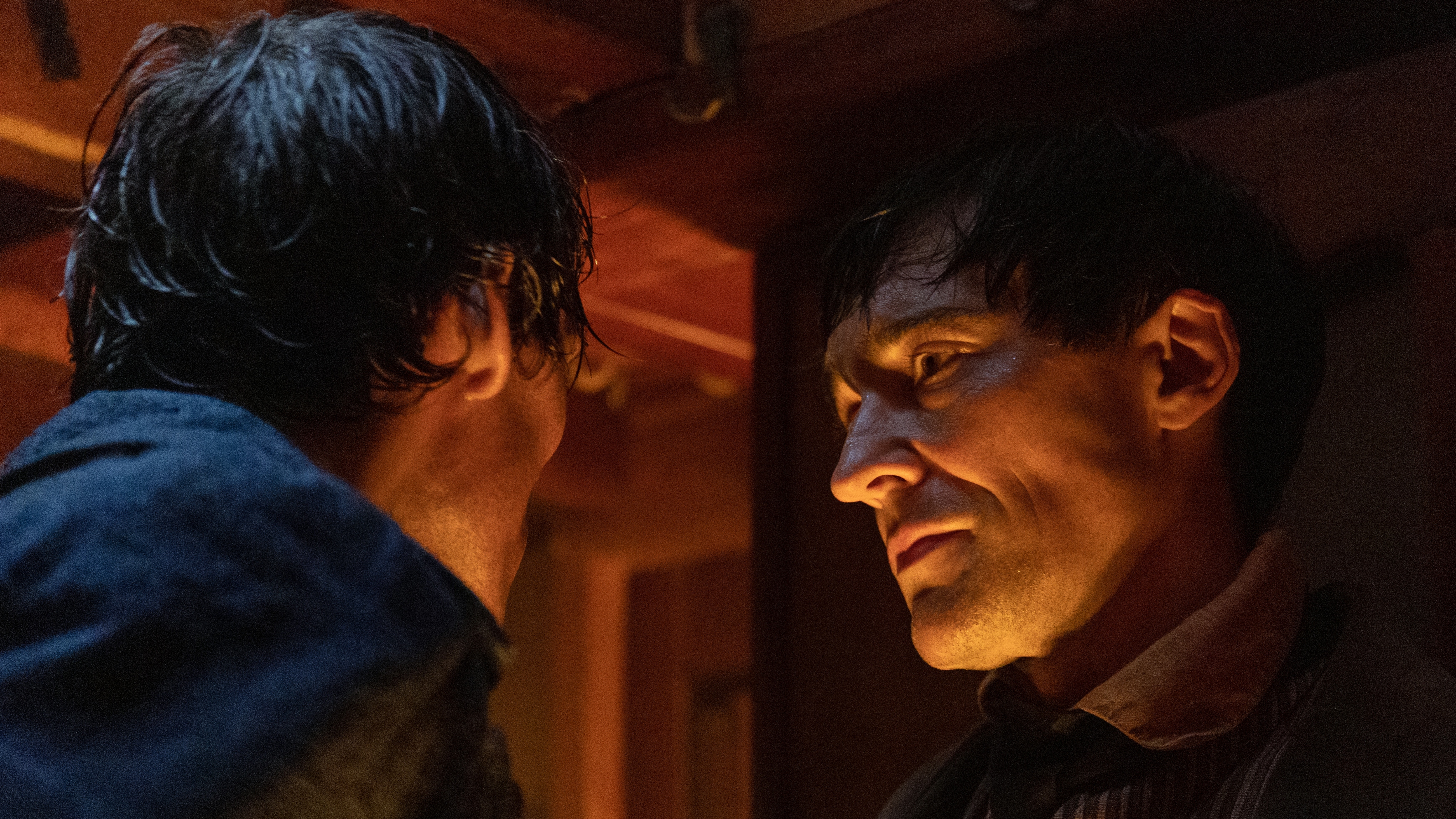 Edmond (Sam Claflin) and Danglars (Blake Ritson) have an intense discussion inside the ship. The room is dimly lit in candlelight, and they are in a shadowy corner. Edmond is facing away from the camera, while Danglars is looking towards Edmond. Danglars' face is lit from underneath, again due to candlelight.