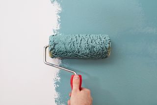 A hand using a paint roller covered in teal paint, rolling it onto a white wall.