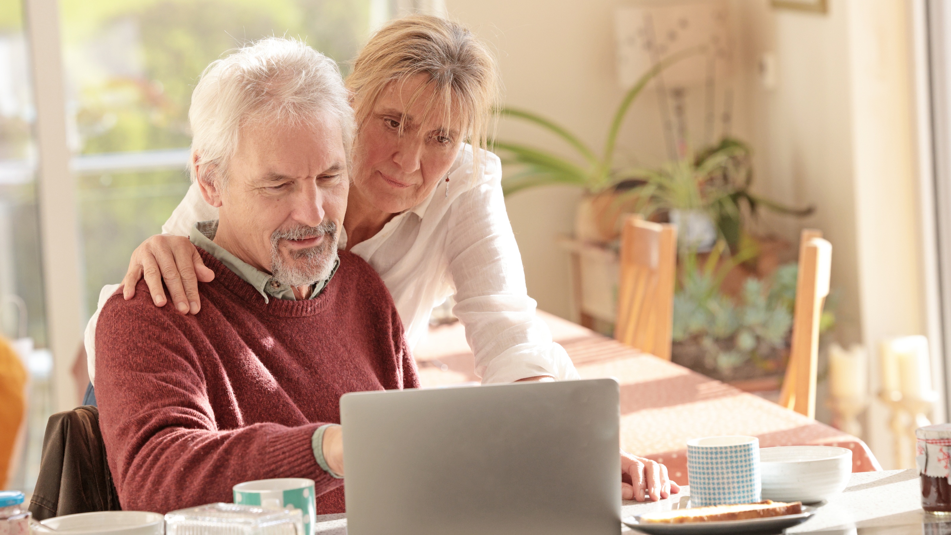 An older couple look at a laptop together at the kitchen table.