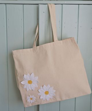 Large crocheted daisy motifs used to decorate a tote bag hanging on a light green cupboard door.
