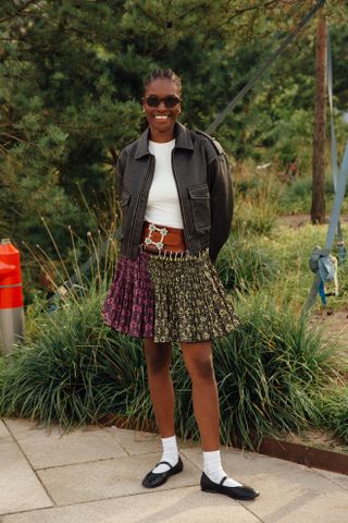 Woman at fashion week wearing preppy outfit featuring a white tee, black leather bomber jacket, plaid mini skirt, white socks, and Mary Jane flats.