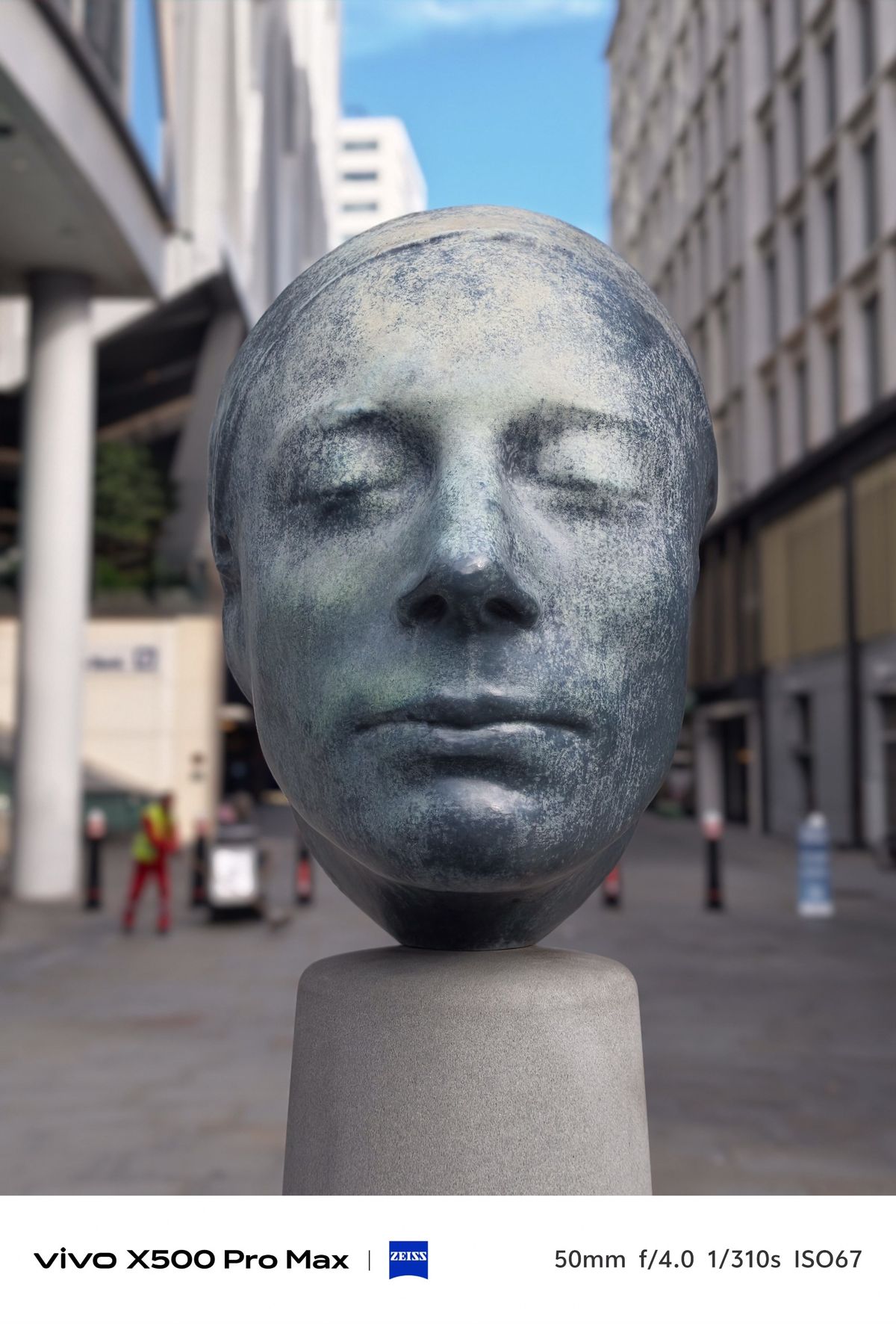 Large sculpted human head with closed eyes displayed on a pedestal between modern city buildings.