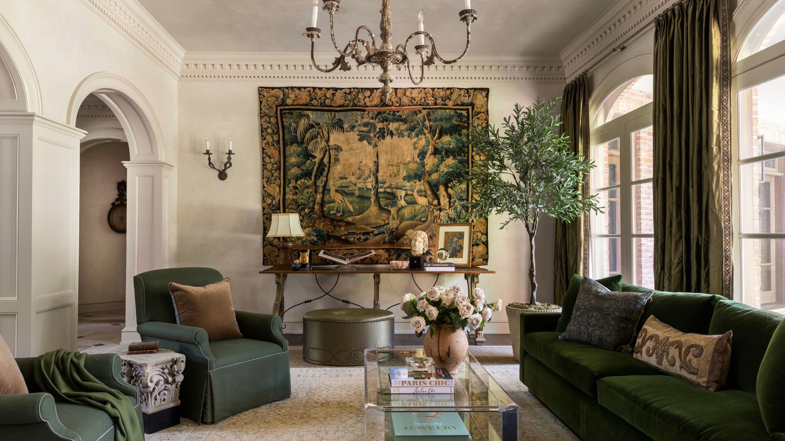 Elegant living room featuring green velvet seating, a large tapestry hanging on the wall, an potted olive tree, and a glass coffee table.