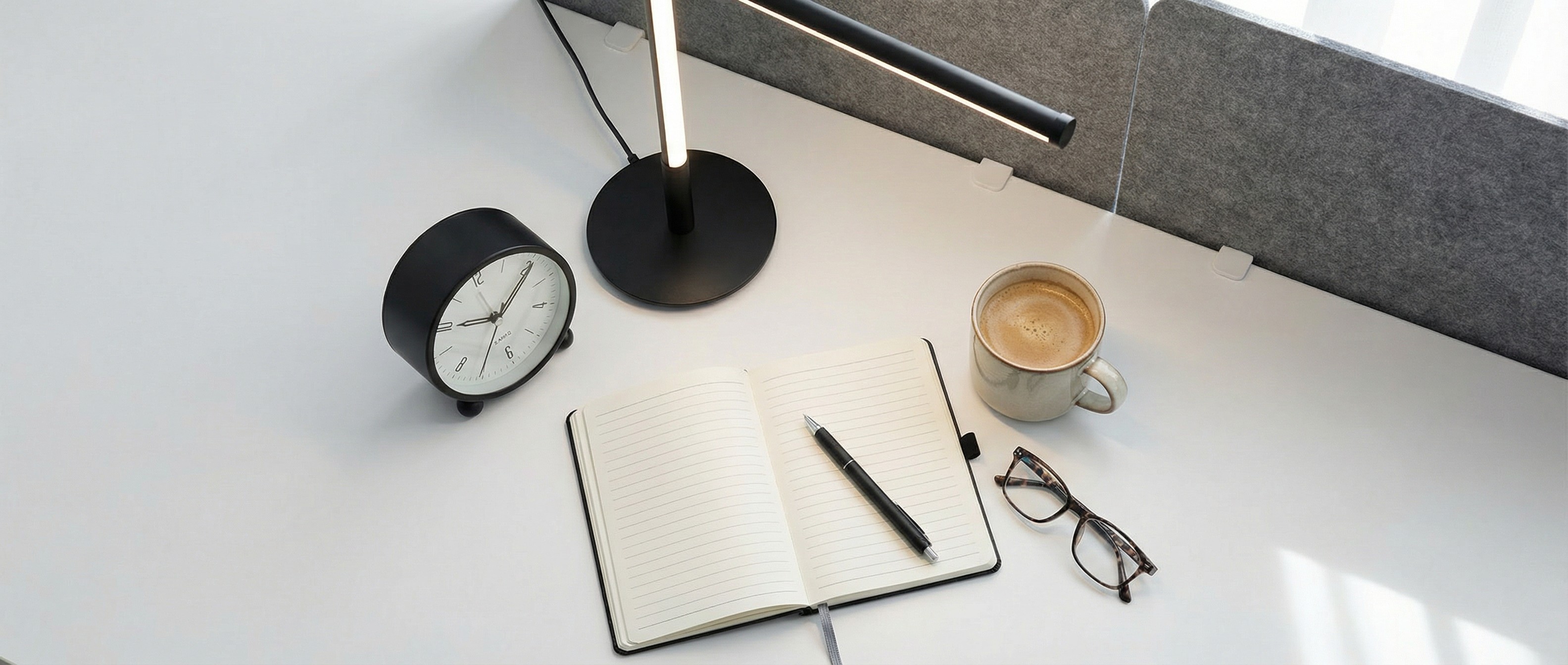 A notebook, lamp, coffee mug, and clock all grey on a desk with a privacy screen