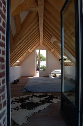 Attic room in VW House by Franklin Azzi, pointed ceiling with wooden beams, white walls, brick wall and door open into the room, low bed on the floor with grey cover, pillows stacked to the side, sky light windows, rug