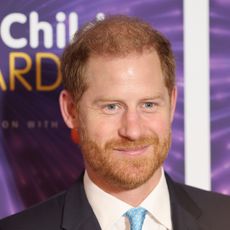 Prince Harry wearing a suit and blue tie standing in front of a purple background