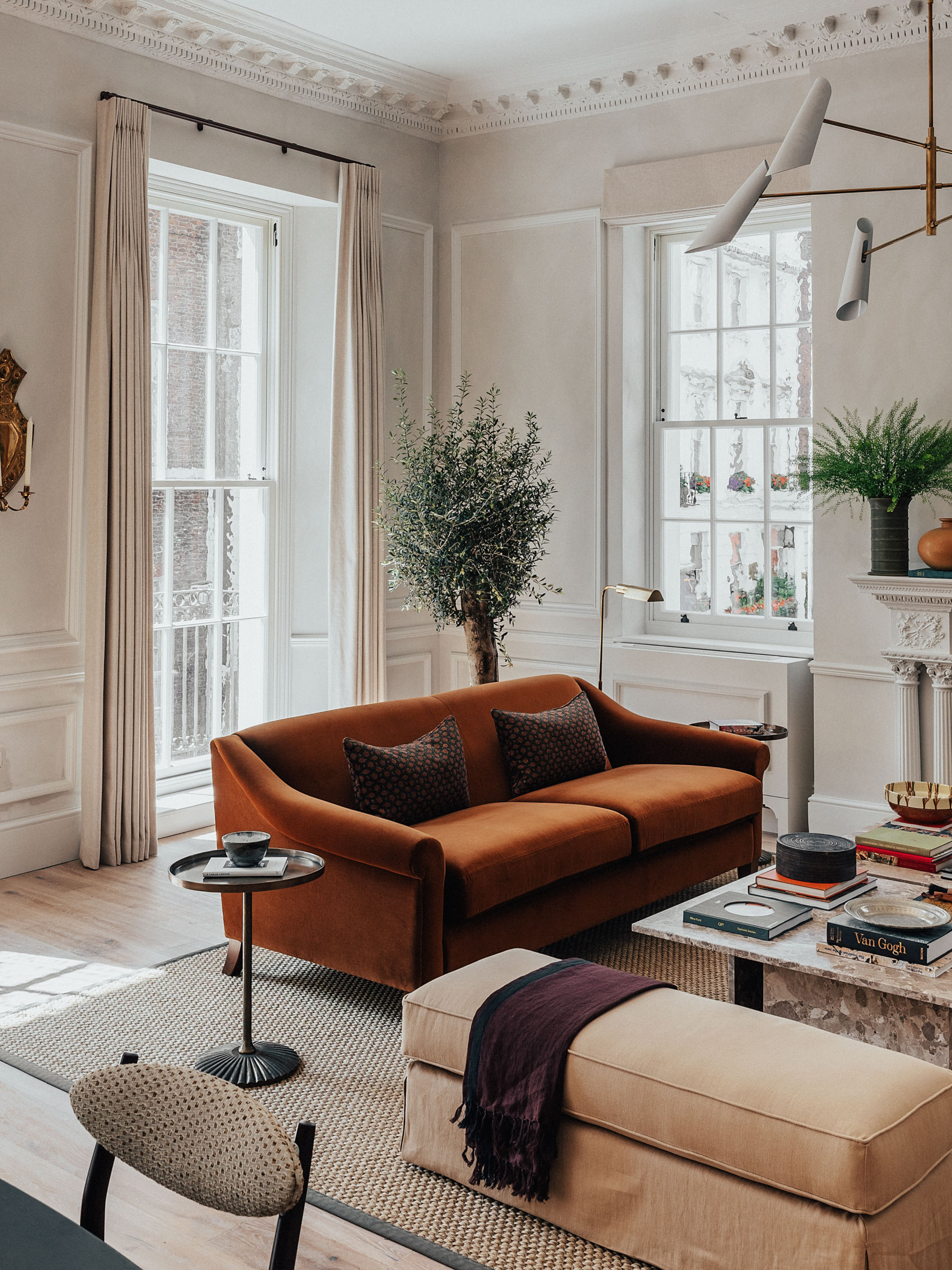 modern living room with orange sofa, long ottoman seat, side table, square marble coffee table styled with books, a potted plant in the corner, sash windows with curtains, decorative cornice, and a jute rug