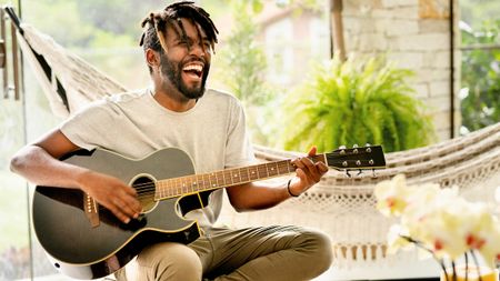 Man smiling whilst playing a black acoustic guitar in front of a hammock