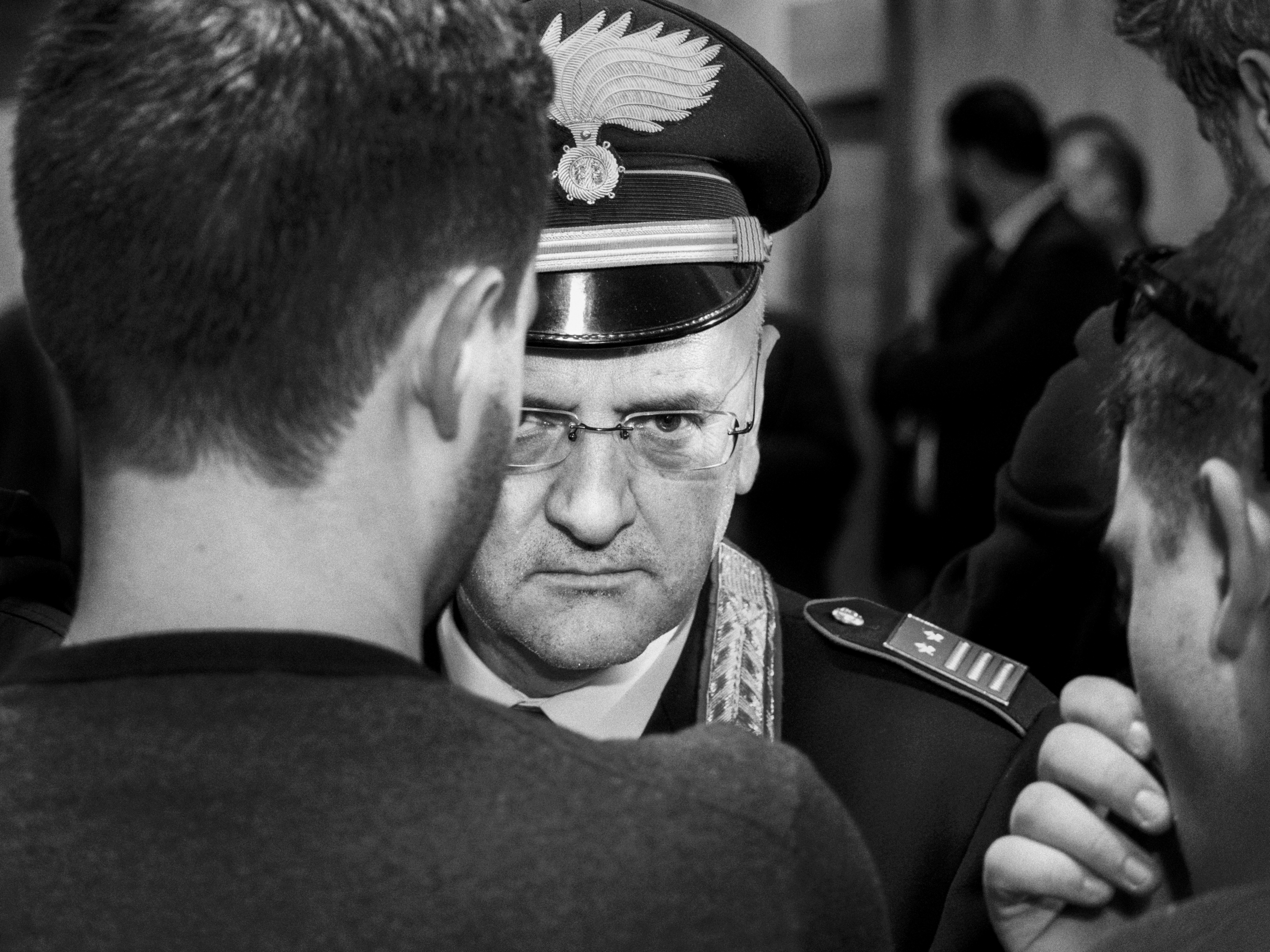 A black-and-white, close-up portrait of an Italian Carabinieri officer in uniform, looking sternly directly at the camera through wire-rimmed glasses between two blurred figures in the foreground.