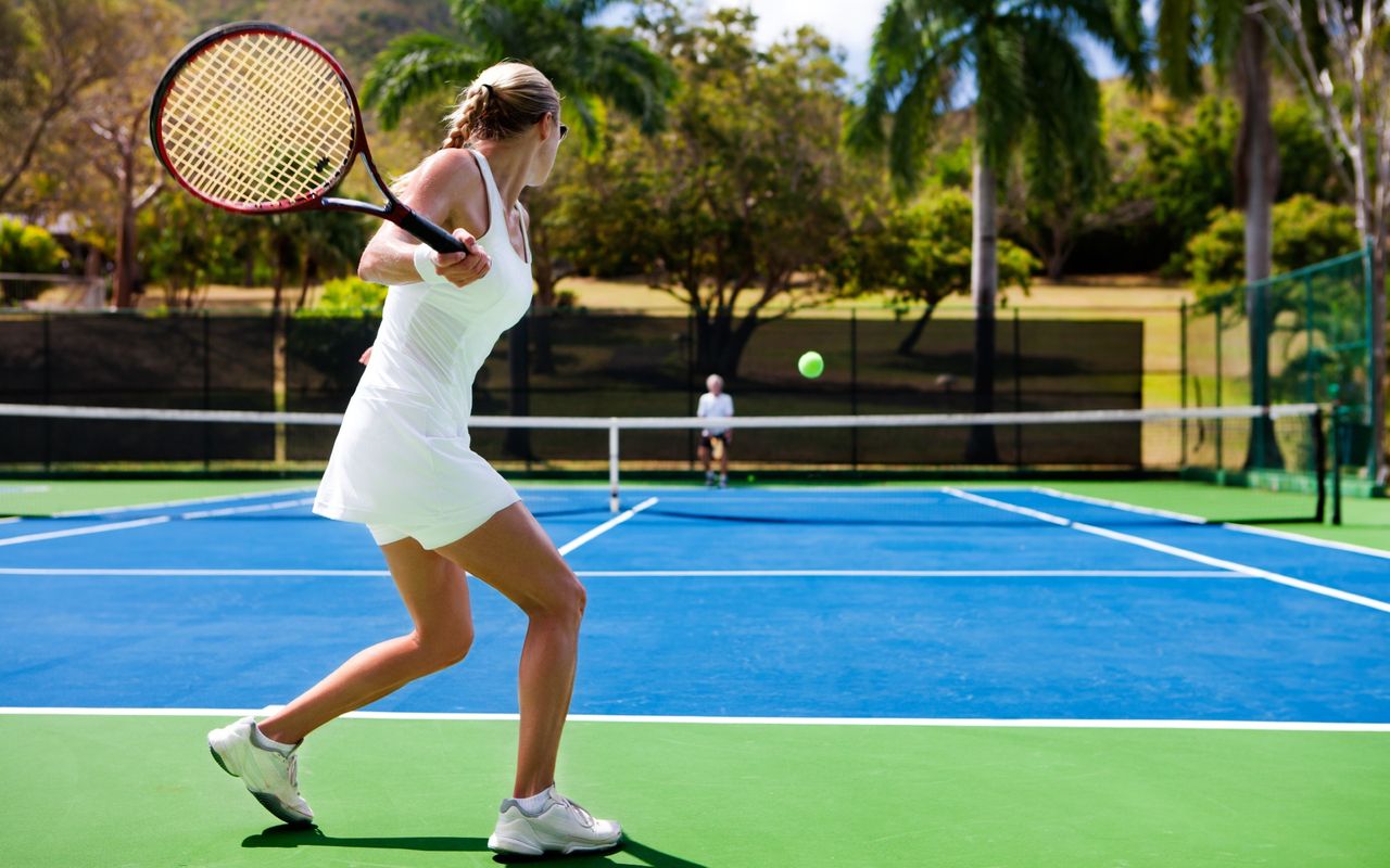 A woman with her blonde hair in a braid prepares to hit a tennis ball across the net at her playing partner who is an older man with white hair