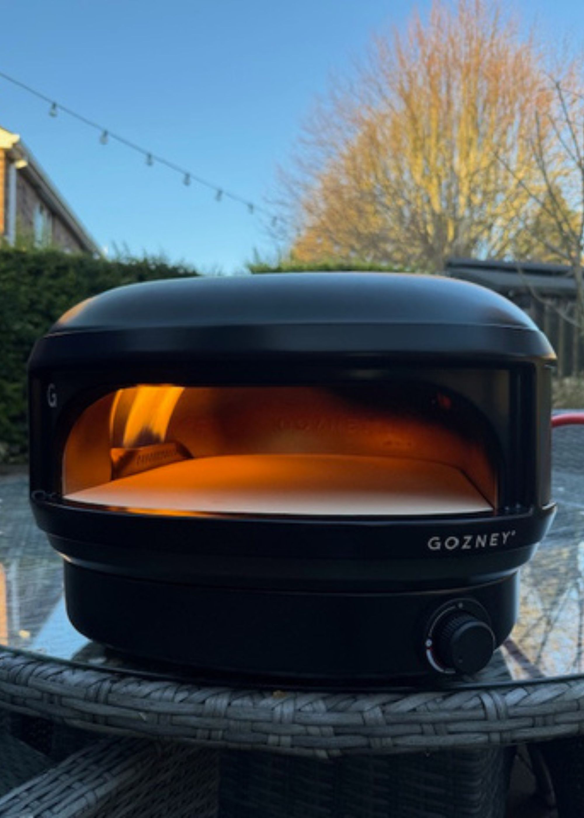 Gozney's Arc Lite pizza oven in off black sitting on top of a round glass-topped rattan garden table outdoors with a tree and blue sky in the background