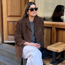 British style influencer Anna Newton poses on a bench outside a cafe in London wearing black Ray Ban wayfarer sunglasses, a brown suede blazer, a gray crewneck sweater, and white jeans