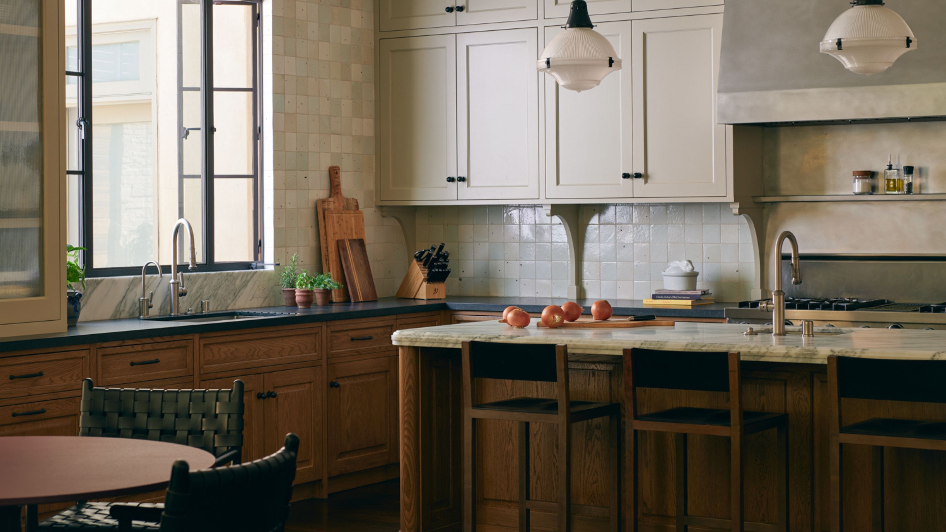 a rustic oak kitchen with two tone upper cream cabinets, zellige tiles, a stainless steel cooker hood and an oak island with bar chairs
