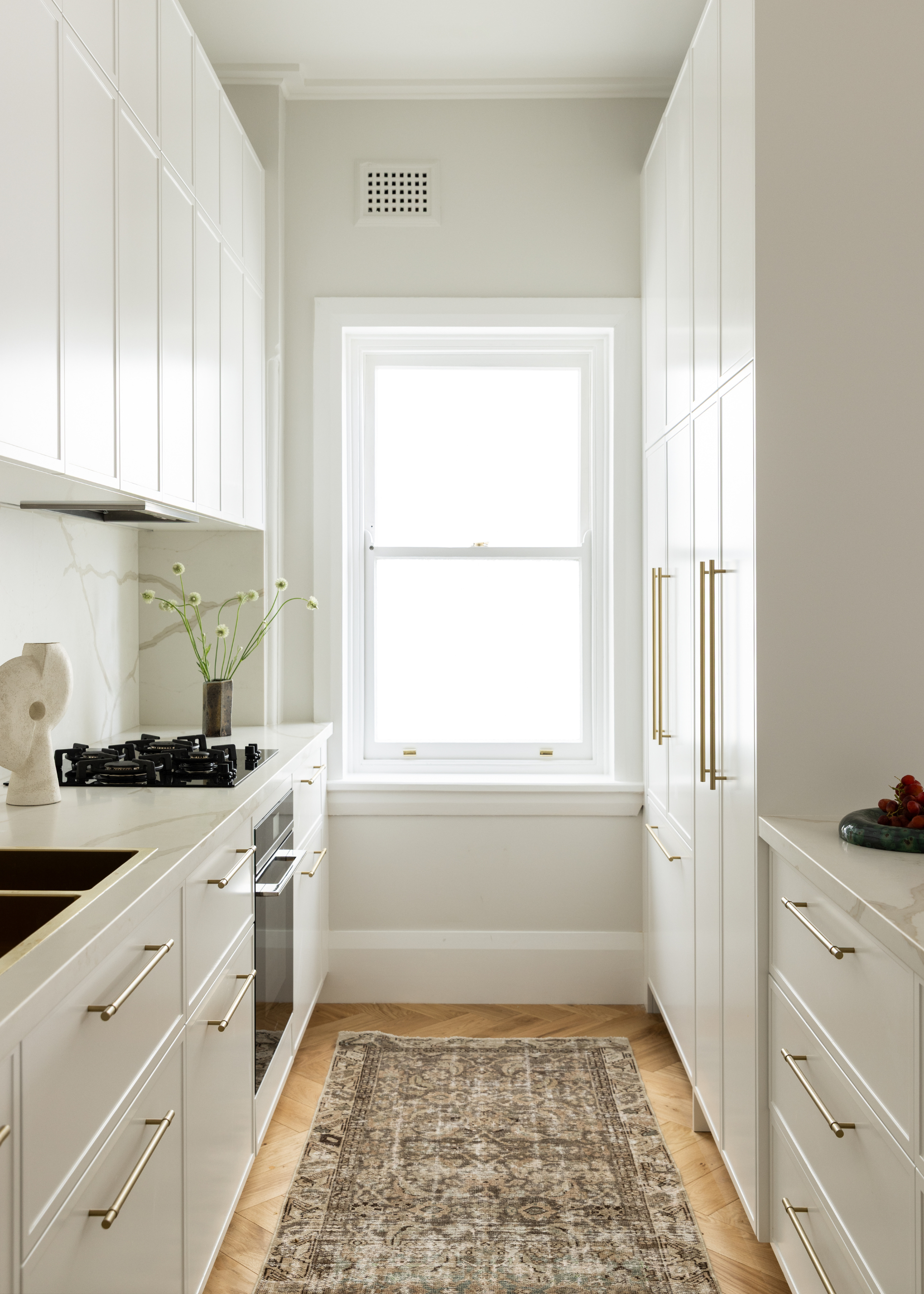 A narrow white kitchen with a gas stove, a vase with flowers, silver hardware on cabinets, and a runner on the floor
