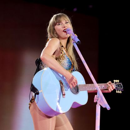 a photo of a white woman with blonde hair using a blue guitar and singing into a purple microphone at a concert