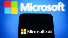 A smartphone with the Microsoft 365 logo displayed, held in front of a blurred Microsoft banner