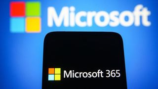 A smartphone with the Microsoft 365 logo displayed, held in front of a blurred Microsoft banner