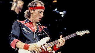 Mark Knopfler performing live onstage, playing Schecter-Suhr Strat guitar, wearing headband 
