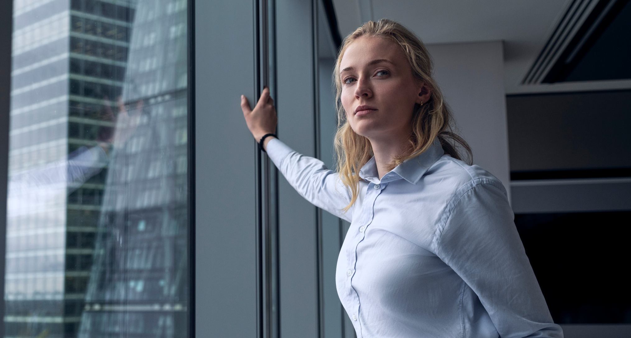 sophie turner as zara wearing a light blue button down leaning against a window in an office building in a still from steal