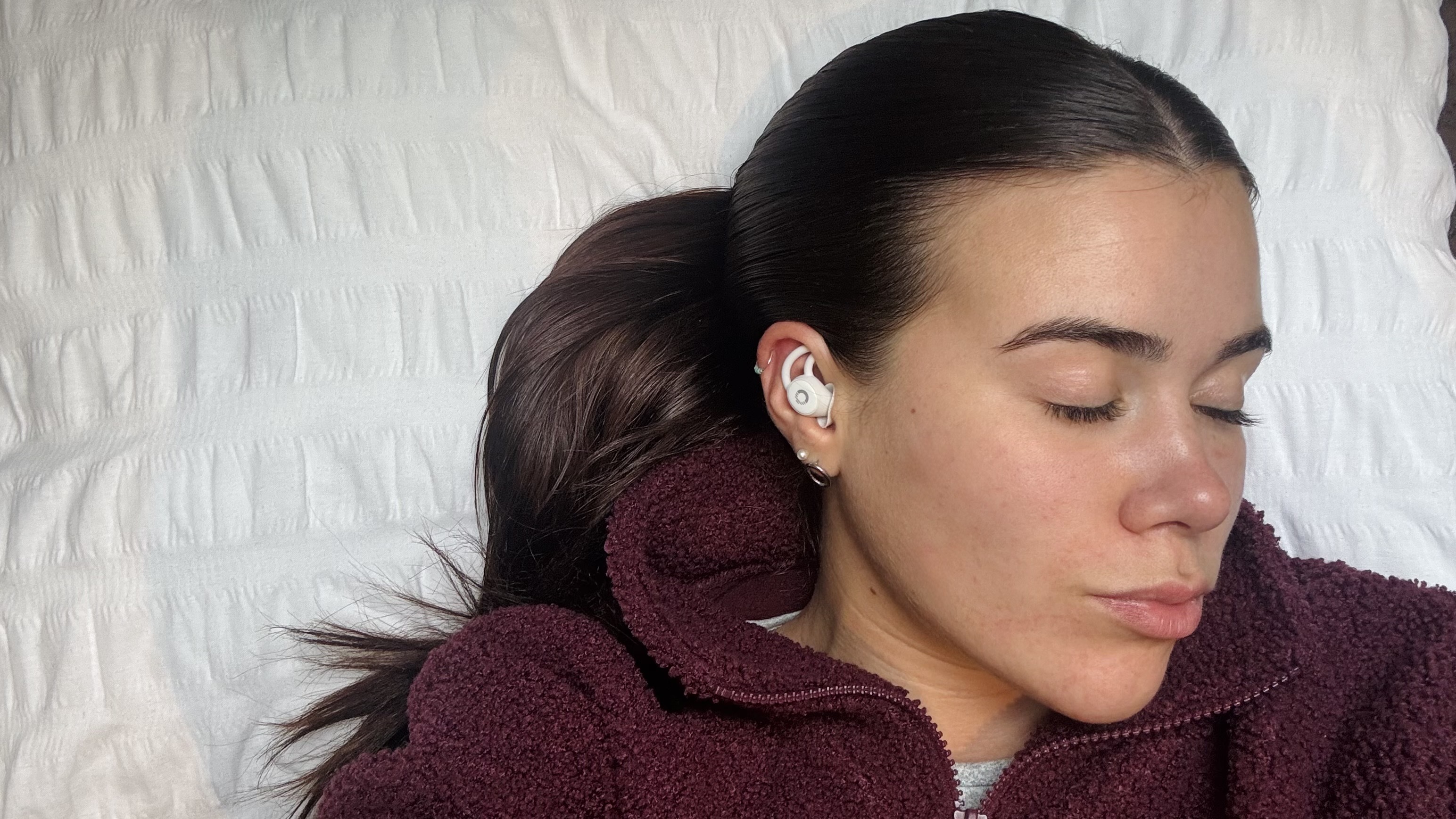 Our sleep tech tester laying on a white pillow wearing a purple jumper and the Ozlo Sleepbuds in her ear