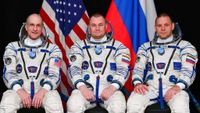 three astronauts in spacesuits sitting down in front of an american flag and a russian flag