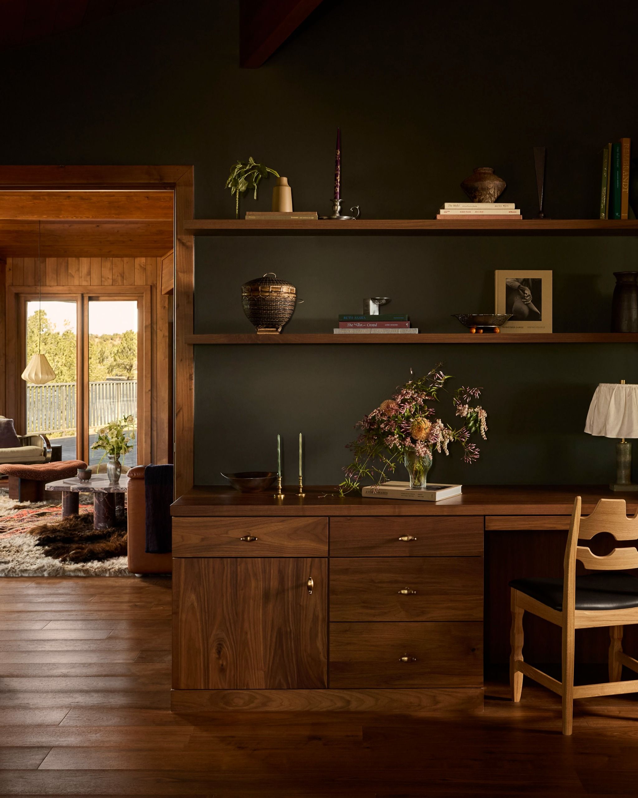 A home office space with a built-in wooden desk and floating wood shelves, dark green wall paint, and a number of decorative items on the desk and shelves with the living room in view through the door