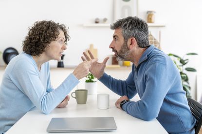 Frustrated mature couple confronting each other, discussing relationship tensions while seated in contemporary kitchen setting, displaying emotional strain