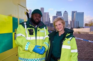 Bloods season 2: paramedics Maleek (Samson Kayo) and Wendy (Jane Horrocks) stand next to the ambulance in their uniforms and bright yellow hi-vis jackets. Behind them is some police tape cordoning off the scene of an accident, and in the far distance behind them is the skyline of Canary Wharf with all its skyscrapers