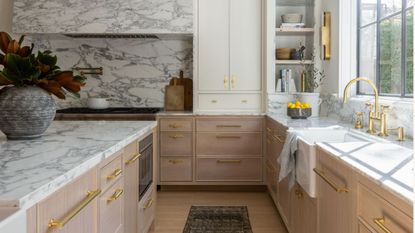 A large kitchen with wooden lower cabinets and white painted upper cabinst, gold hardware and taps, a large kitchen island and a marble backsplash