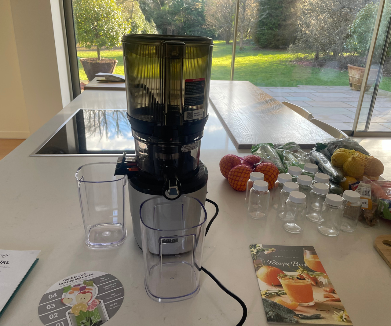 A black and stainless steel Kuvings Auto10 Hands-Free Juicer is on the counter in our bright test kitchen. There is also fruit, vegetables, and instruction manuals in view.