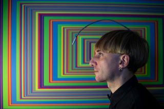 A photo of Neil Harbisson, who hears color through an antenna implanted in his skull, next to a piece of his artwork.