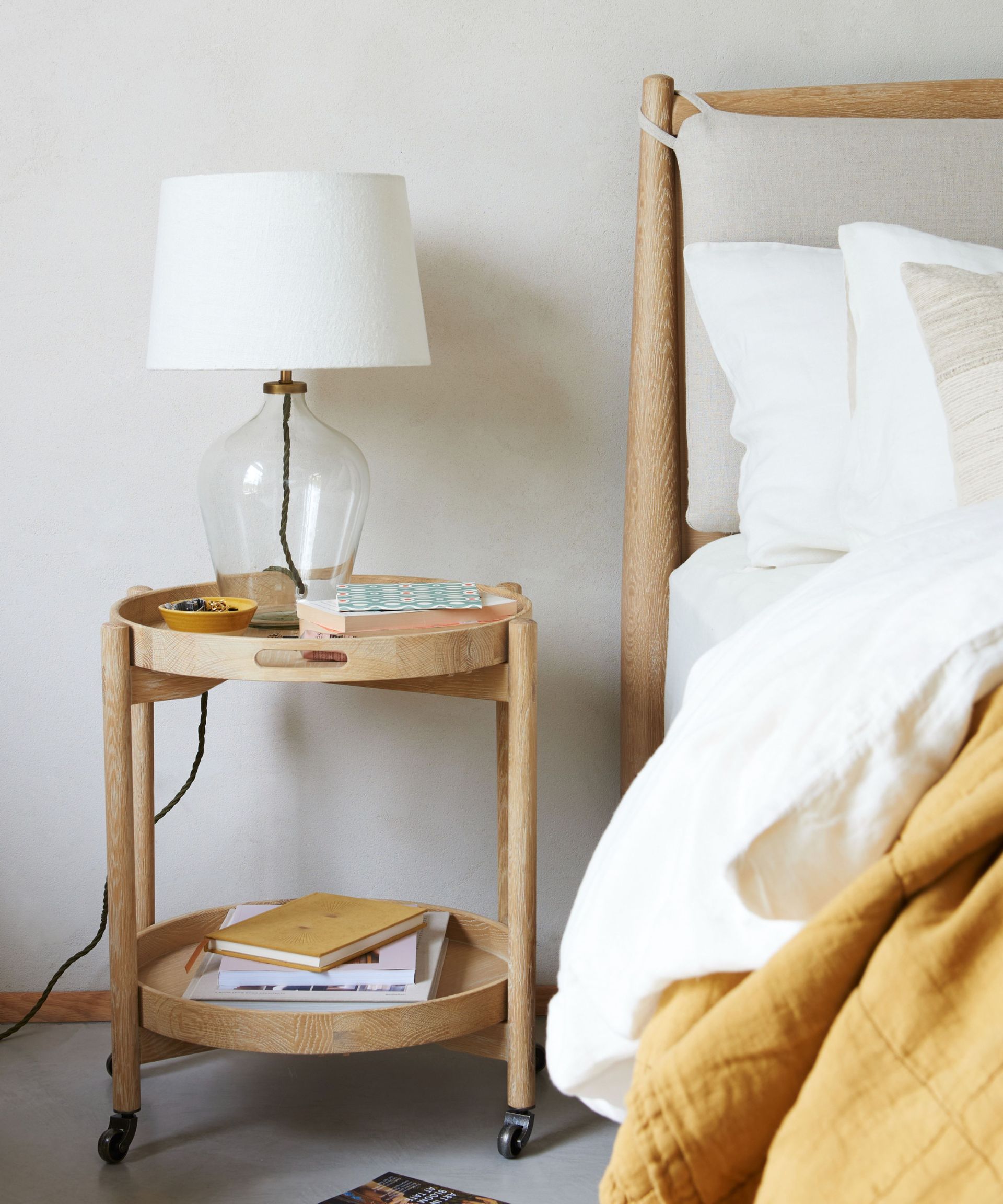 neutral bedroom with wooden bed with white and yellow bedding, matching nightstand with legs, clear glass table lamp base with white shade, books on lower shelf