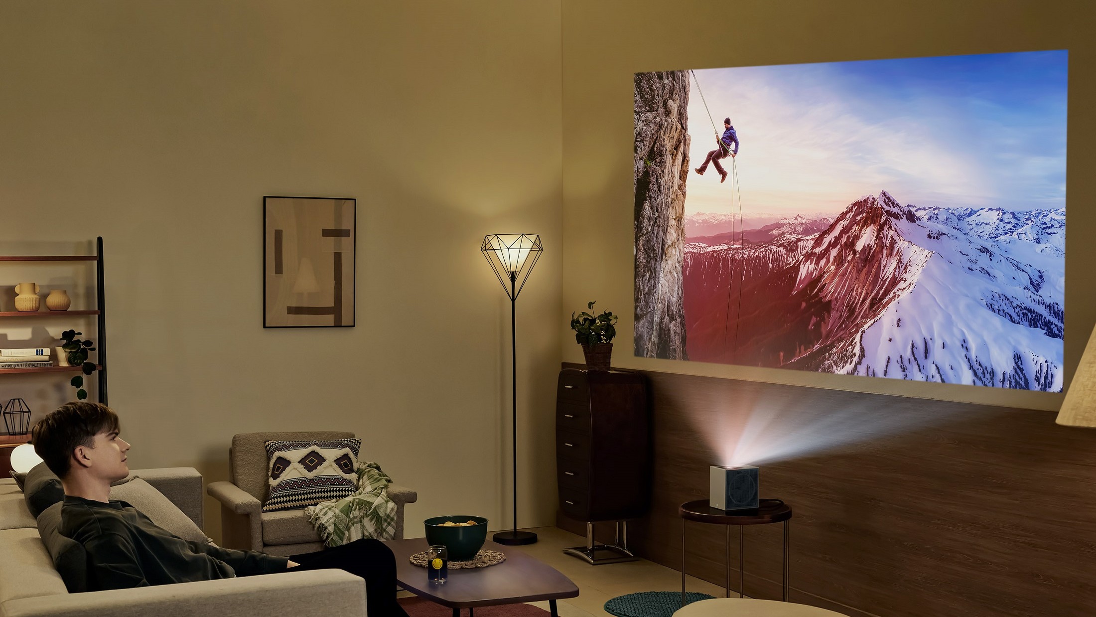 A modern living room with a man watching a projector at the front of the room. A climber is on screen descending down a cliff face.