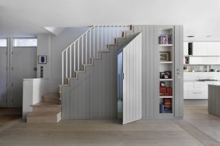 A small utility room behind a grey built in door under a staircase beside white kitchen