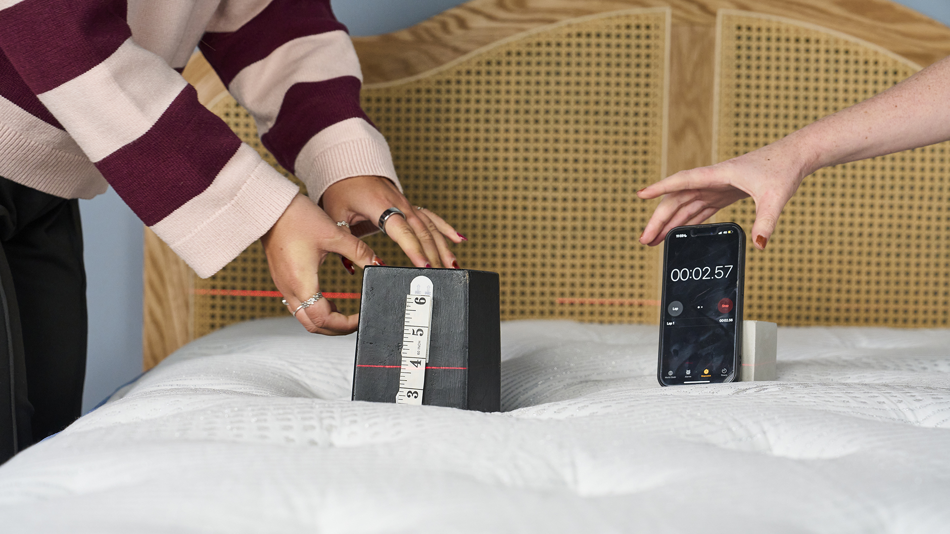 Two pairs of hands are shown on the Bear Elite Hybrid mattress. One pair is holding a black rectangular weight 25kg with a measuring tape attached, while the other pair is holding a smartphone displaying a stopwatch reading of 00:02.57. A red laser line is projected across the weight to indicate how deep it sinks into the mattress surface.
