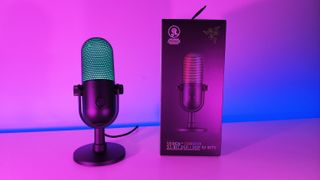 Razer Seiren V3 Chroma microphone illuminated in cyan RGB next to its retail packaging box under neon lighting.