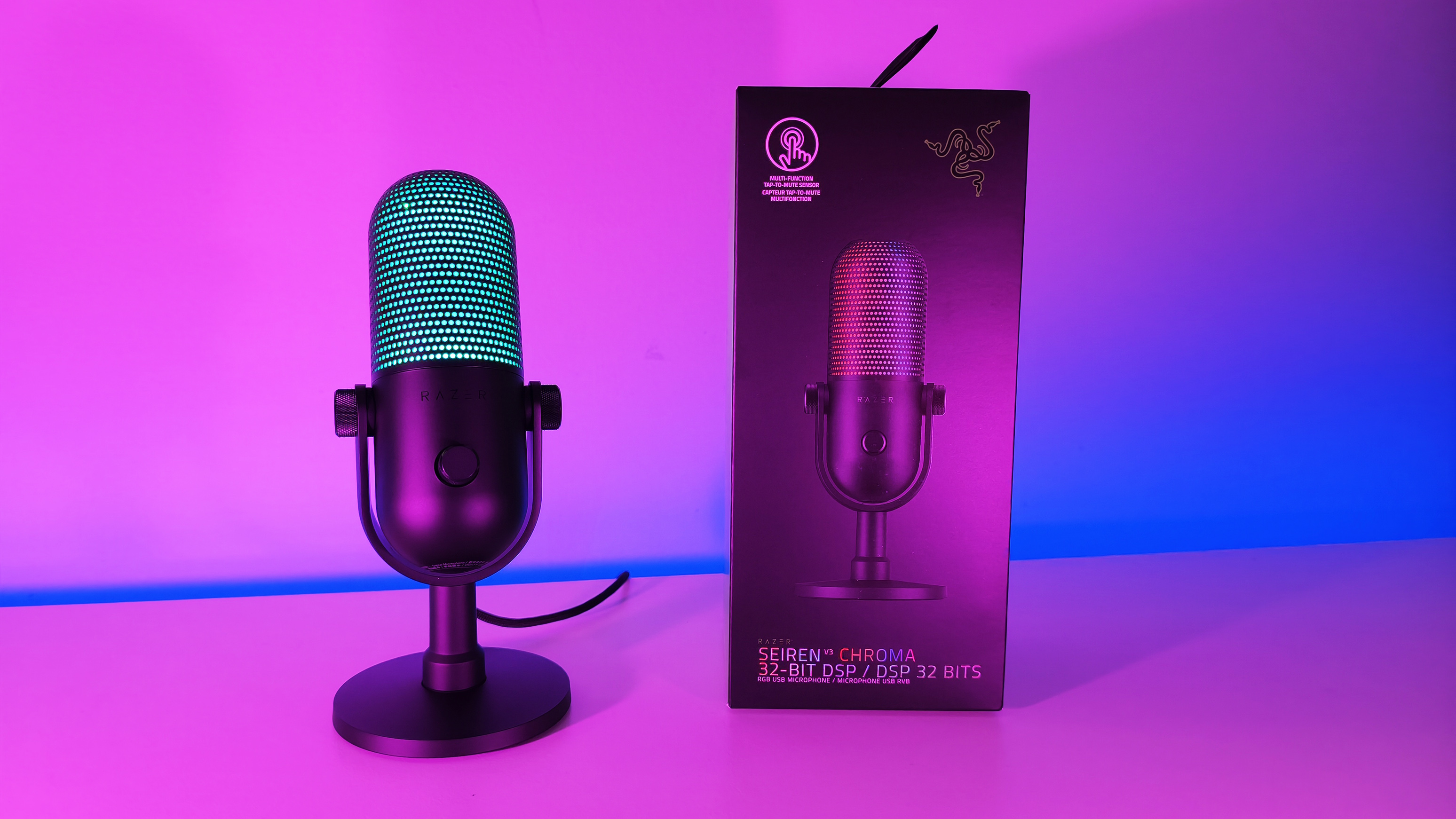 Razer Seiren V3 Chroma microphone illuminated in cyan RGB next to its retail packaging box under neon lighting.