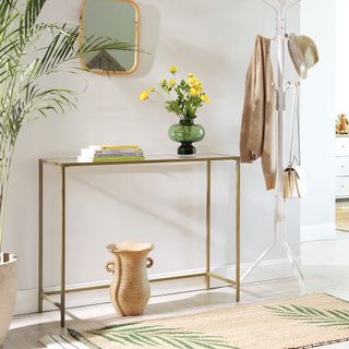 a slick modern gold and glass console table with a hanging rack next to it and a pot below it, and coffee table books and flowers on top