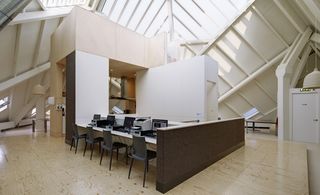 Alternative interior view of the Super Cube featuring white, brown and wood covered walls, light coloured flooring, a pendant light, a long desk with computers and a printer on top and four dark grey chairs
