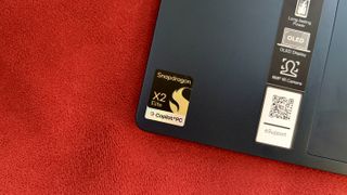 Corner of a laptop on a red fabric surface, featuring a Snapdragon X2 Elite sticker, indicating the use of a powerful processor and Copilot+PC technology.