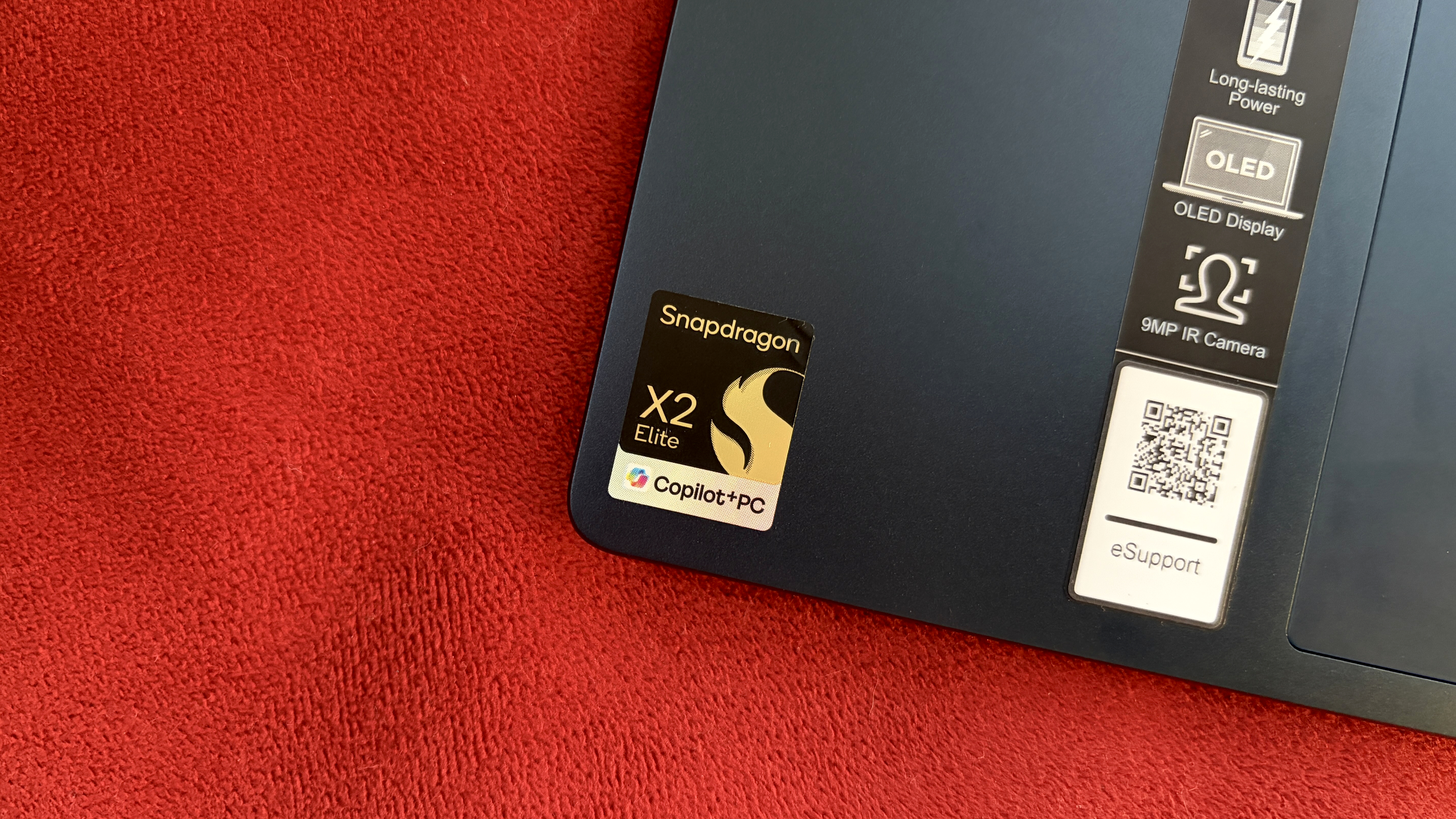 Corner of a laptop on a red fabric surface, featuring a Snapdragon X2 Elite sticker, indicating the use of a powerful processor and Copilot+PC technology.